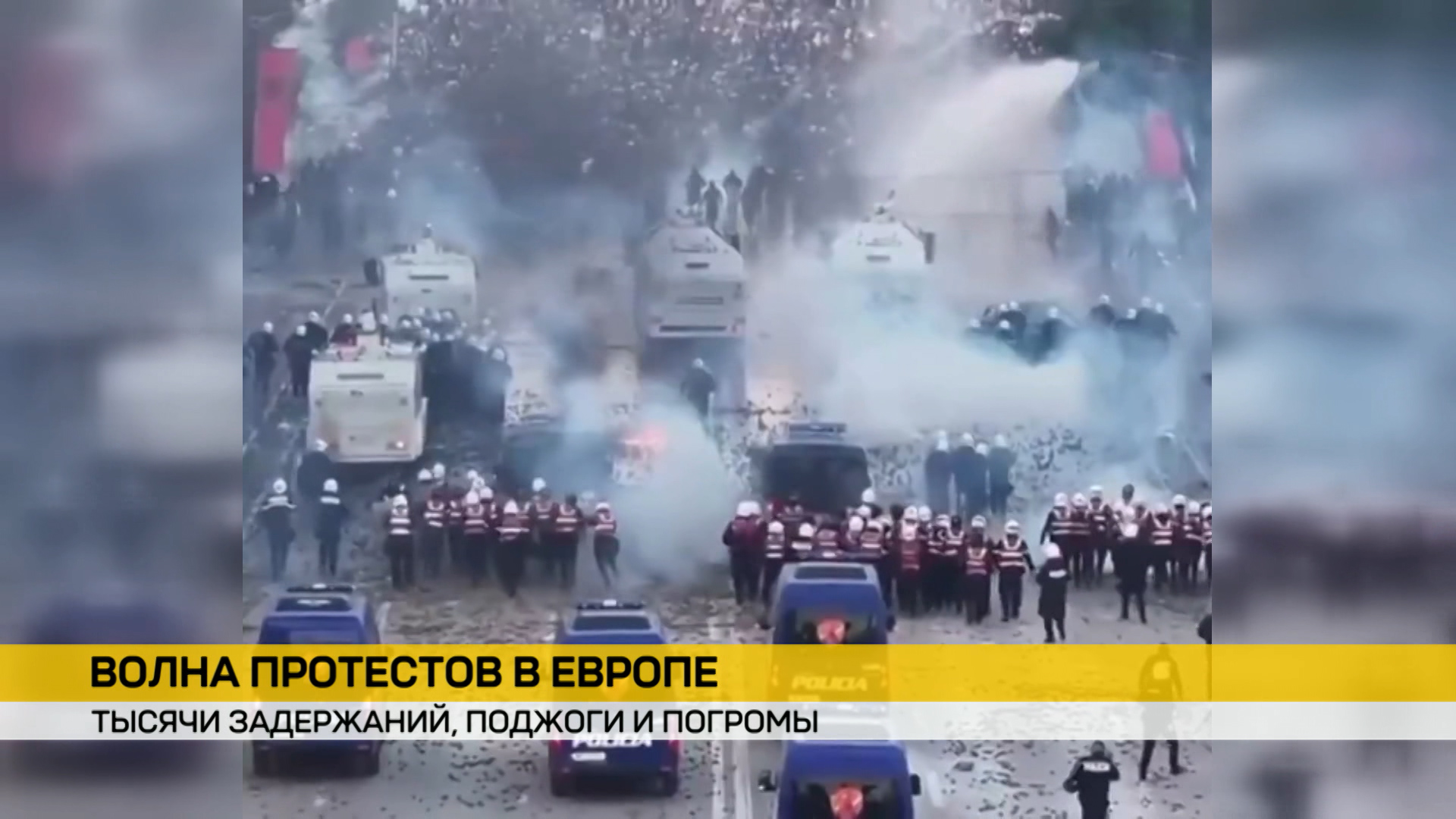 Столкновения протестующих и полиции на улице европейского города. Спецтехника использует водометы, вокруг дым, силовики выстроены в шеренги.